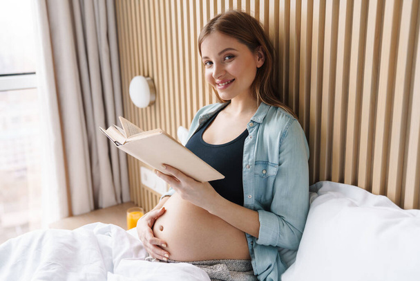 Photo of happy pregnant woman smiling and reading book while sitting in bed at sunny room - Fotó, kép