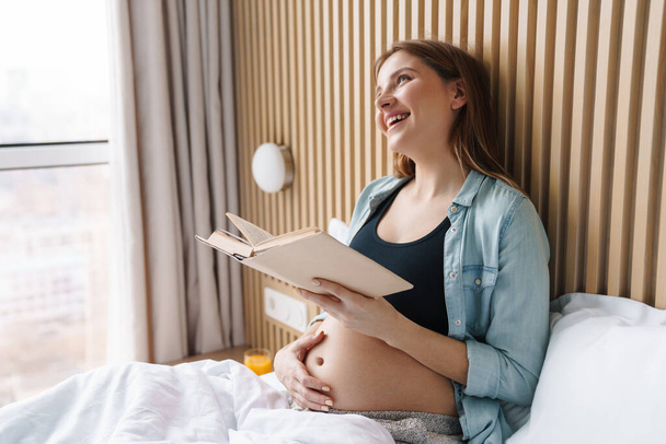 Photo of happy pregnant woman smiling and reading book while sitting in bed at sunny room - 写真・画像