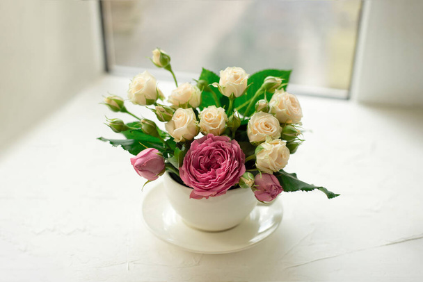 A bouquet of white roses in a cup on a white table opposite the window - Foto, Imagen