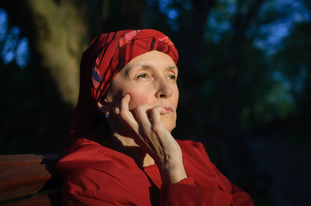 Female portrait of mature woman dressed in red clothes and sitting on the bench and enjoying a good weather and sunset in the park during a spring or autumn. - Photo, Image