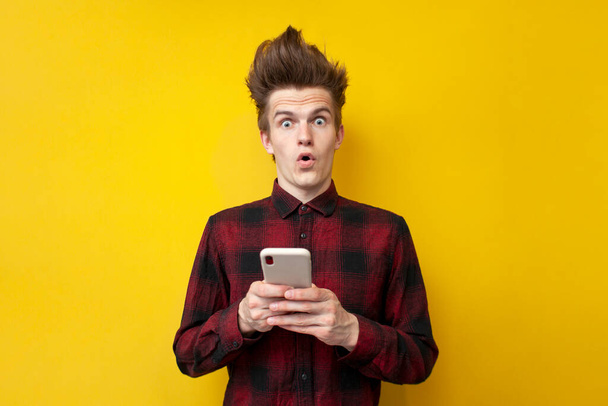 young shocked guy with funny hairstyle use smartphone on yellow isolated background, hipster holds phone and is surprised - Photo, Image