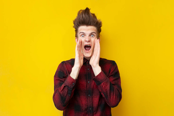 νεαρός σοκαρισμένος τύπος ουρλιάζοντας σε ένα κίτρινο απομονωμένο φόντο, hipster με ένα αστείο χτένισμα είναι πανικός, γκρο πλαν - Φωτογραφία, εικόνα