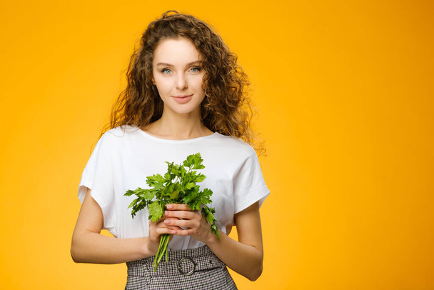 Portrait of pretty caucasian girl with curly hair holding parsley isolated on colorful yellow background in studio - Foto, afbeelding