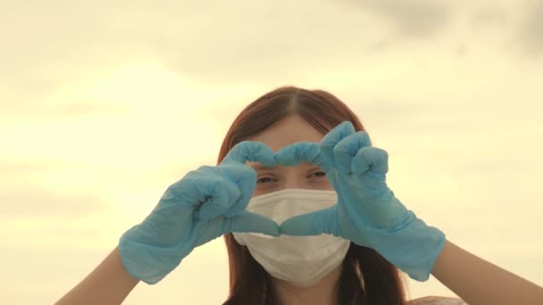 κορίτσι με προστατευτική μάσκα και ιατρικά γάντια δείχνει το σύμβολο της καρδιάς με τα χέρια της. Η νεαρή γυναίκα αγαπάει τους γιατρούς. πανδημία, covid 19. γυναίκα γιατρός δείχνει δάχτυλα καρδιάς και την αγάπη για τους ασθενείς. - Πλάνα, βίντεο