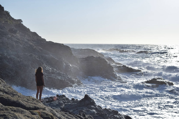 Дівчинка дивиться драматургічно на кругозір Егейського моря, а хвилі кружляють навколо неї в Ікарії, Греція. - Фото, зображення