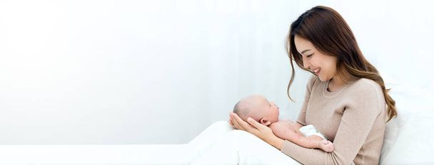 Όμορφη μαμά αγκαλιάζει απαλά το νεογέννητο μωρό, ενώ το βρέφος κοιμάται στην αγκαλιά. Ασιάτισσα μητέρα κοιτάζει το μωρό με αγάπη και δείχνει προστασία. - Φωτογραφία, εικόνα