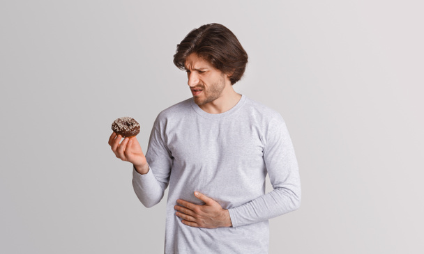 Мужчина держит сладкий пончик в руках и смотрит на него с болью, сжимая руки к животу
 - Фото, изображение