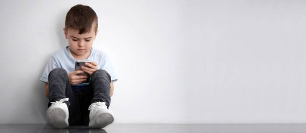Sad little boy with mobile phone sitting near white wall, banner design and space for text. Time to visit child psychologist - Foto, imagen