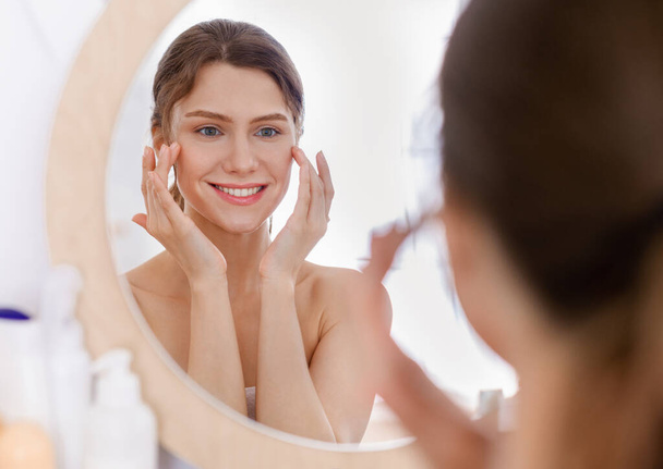 Красивая женщина смотрит в зеркало и применяет глазную сыворотку
 - Фото, изображение