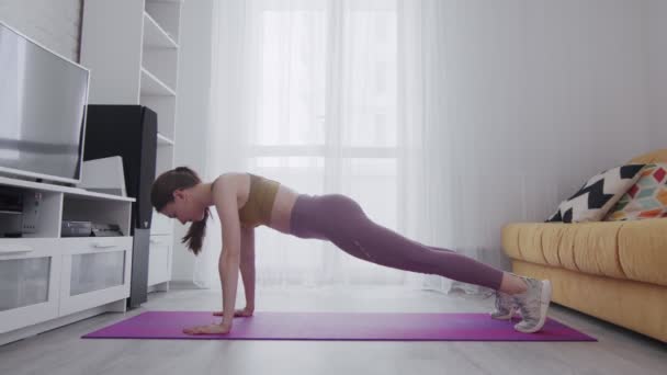 Young beautiful woman with tattoo doing static plank on yoga mat while training at home - Footage, Video