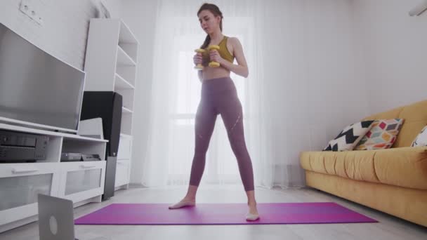 Young beautiful woman does squats with dumbbell with help of his instructor online on laptop at home - Πλάνα, βίντεο