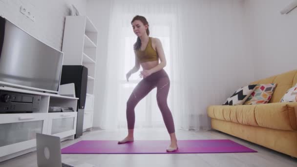 Young beautiful woman does squats with help of his instructor online on laptop at home - Materiał filmowy, wideo