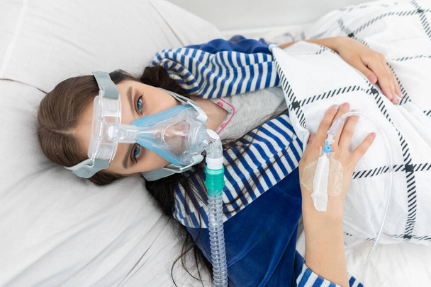 Oxygen mask attached to the respiratory tract. The teenager lies in a hospital bed. A seriously ill young girl. - Fotoğraf, Görsel