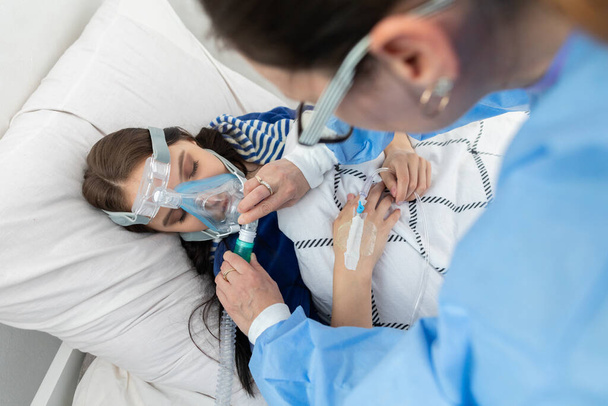 The doctor controls the connection of the ventilator. The teenager lies on a hospital bed. Oxygen mask on the respiratory tract. - Fotoğraf, Görsel