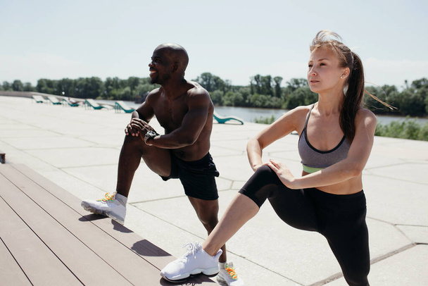 White girl and black man do stretching on the inner muscles of the thighs with his foot resting on a bench - Foto, Imagen