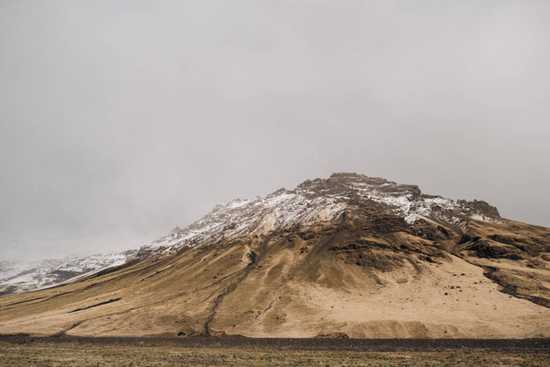 Monte rochoso de pico coberto de neve coberto com grama seca amarela na Islândia
. - Foto, Imagem