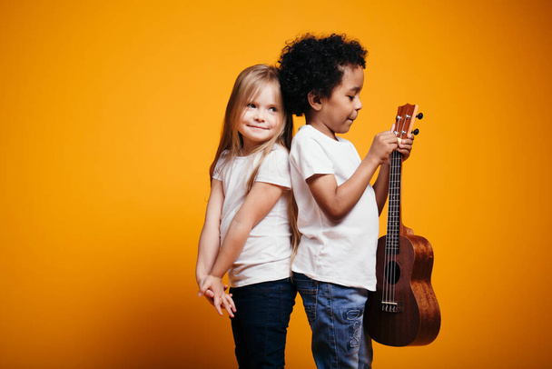 Blonde girl snuggles her back against a dark-skinned boy holding a ukulele guitar in his studio - Fotó, kép