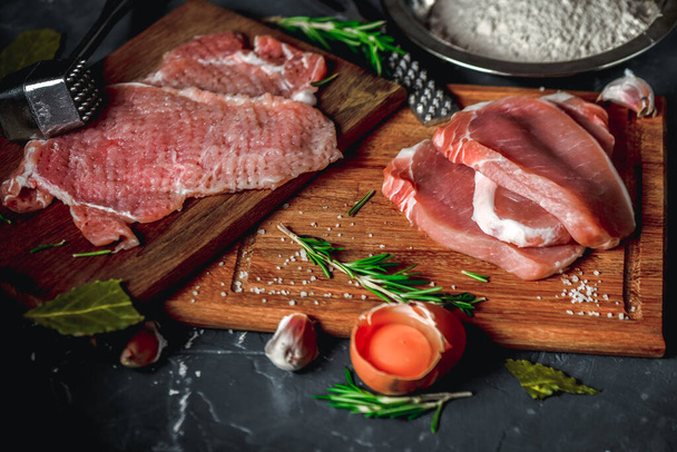 Raw chops and pieces of pork for escalopes on cutting boards with flour, rosemary, bay leaf, sauce on the kitchen table. One piece of meat fell from the chef's hands into flour. Place for text - 写真・画像