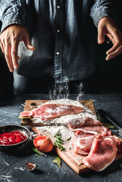 Raw chops and pieces of pork for escalopes on cutting boards with flour, rosemary, bay leaf, sauce on the kitchen table. One piece of meat fell from the chef's hands into flour. Place for text - Fotó, kép