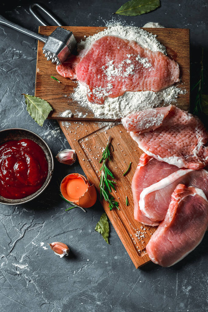Raw chops and pieces of pork for escalopes on cutting boards with flour, rosemary, bay leaf, sauce on the kitchen table. One piece of meat fell from the chef's hands into flour. Place for text - Foto, Imagem