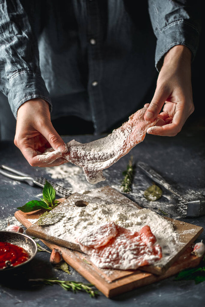 Raw chops and pieces of pork for escalopes on cutting boards with flour, rosemary, bay leaf, sauce on the kitchen table. One piece of meat fell from the chef's hands into flour. Place for text - 写真・画像