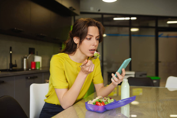 Succesvolle jonge vrouw eten kijken naar smartphone - Foto, afbeelding