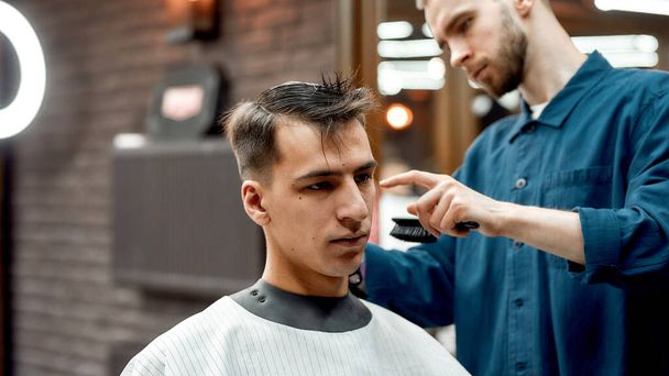 Young handsome guy sitting in armchair and getting haircut at barbershop. Barber working with client, holding hair brush - Foto, afbeelding
