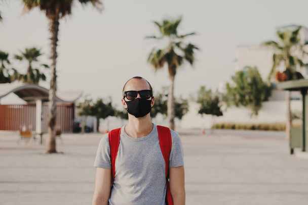 Fiatal férfi utazó hátizsákkal és napszemüvegben fekete védőmaszkot visel. Covid 19 vagy a koronavírus világjárvány koncepciója. Porvédő maszkos férfi, újrahasznosítható - Fotó, kép