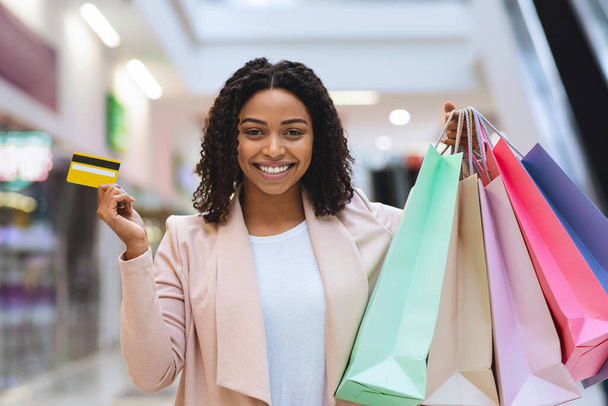 Mujer negra feliz posando con tarjeta de crédito y bolsas de compras en el centro comercial
 - Foto, Imagen