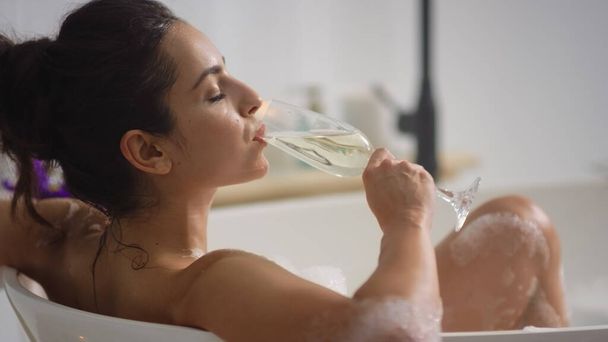 Een close-up van een ontspannen vrouw die wijn drinkt in bad. Romantisch meisje rusten in bad - Foto, afbeelding