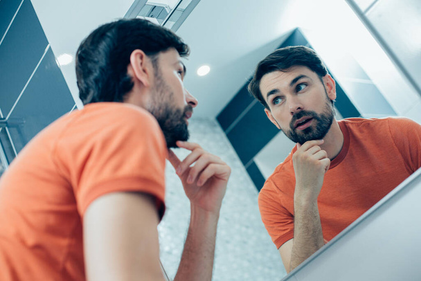 Curieux jeune homme réfléchi debout devant un miroir et touchant son menton tout en regardant le reflet
 - Photo, image