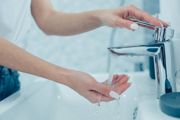 Gros plan d'une femme avec manucure blanche allumant le robinet et mettant une main dans de l'eau courante propre dans le lavabo de la salle de bain
 - Photo, image