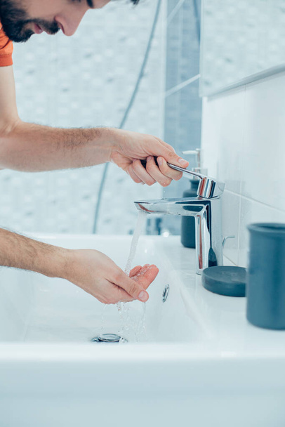 Close up photo of a young man leaning to the bathroom basin and putting his hands under the clean running water - Фото, зображення