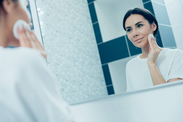 Contented young woman looking at her reflection in the mirror and smiling while touching her cheek with a cotton pad - Foto, Bild