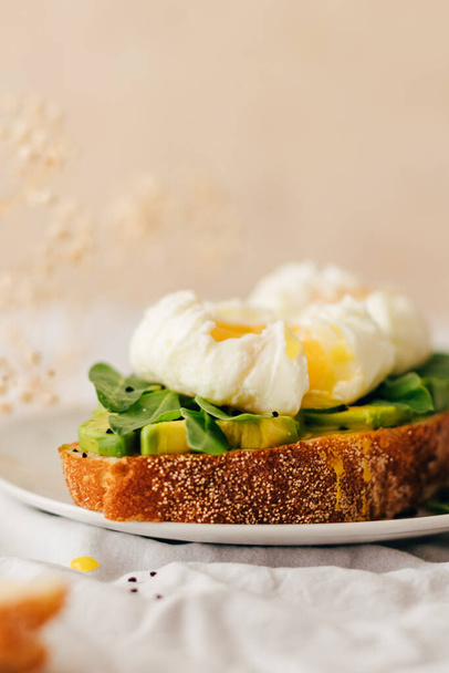 sandwich with whole grain bread, avocado, arugula and poached eggs sprinkled with nigella decorated with small gypsophila flowers on a white plate. textile and beige background. Copy space - Photo, Image