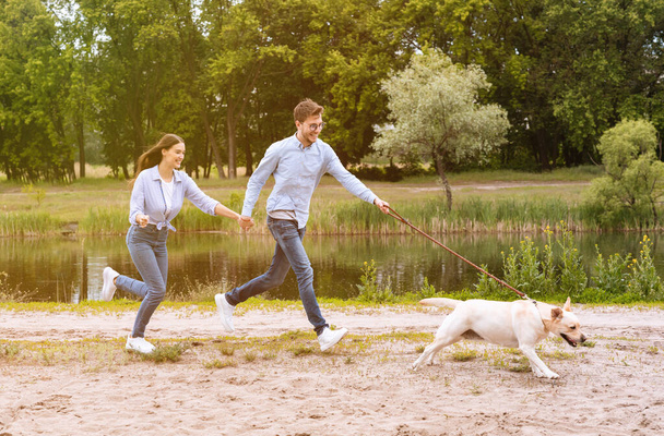 Heyecanlı çift ve av köpeği göl kenarında koşuyor. - Fotoğraf, Görsel