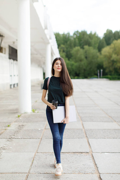 Schöne junge brünette Frau trägt Rucksack und trinkt Kaffee zum Mitnehmen, während sie draußen spaziert - Foto, Bild