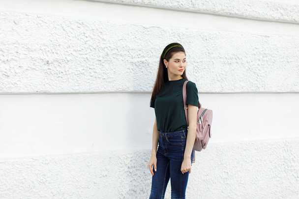 Chica morena en jeans, una camiseta verde, una banda para el pelo y una mochila rosa se levanta contra una pared blanca
 - Foto, Imagen