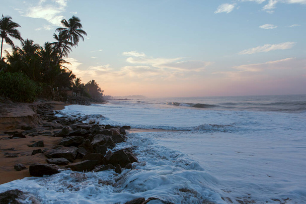 Beautiful postcard landscape shot of the Pitiwella beach (Sri Lanka) at sunrise with orange sand, green palm trees, brown rocks, the Indian ocean with big white waves and the sun on the horizon. - Foto, imagen
