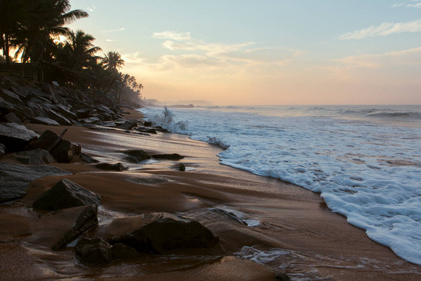 Beautiful postcard landscape shot of the Pitiwella beach (Sri Lanka) on sunrise with orange sand, green palm trees, brown rocks, the Indian ocean with big white waves and the sun on the horizon. - Fotoğraf, Görsel