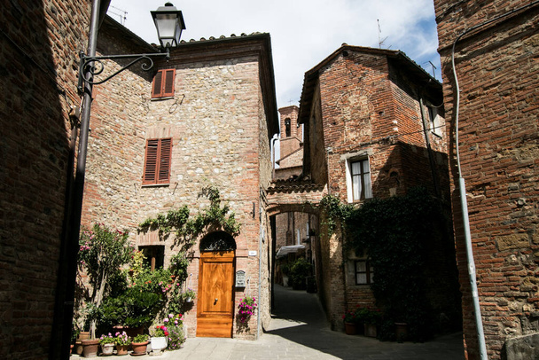 Маленькая площадь средневековой итальянской деревни, с кирпичными домами, деревянной дверью и зелеными и цветущими растениями. Путешествие по Италии
 - Фото, изображение