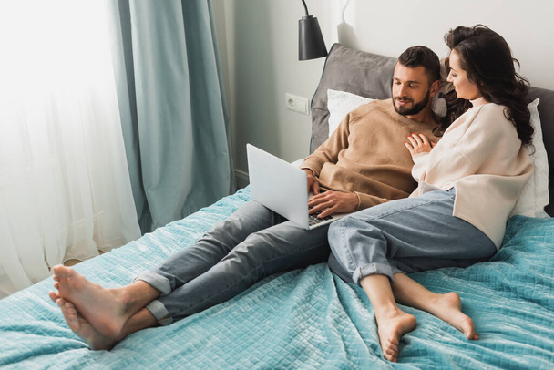 happy freelancer using laptop near beautiful girlfriend in bedroom  - Foto, afbeelding