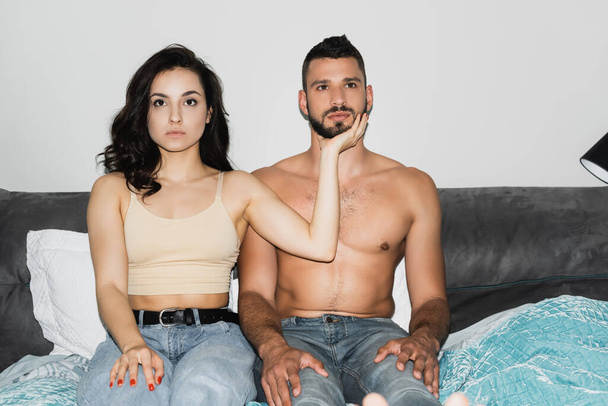 ベッドルームに座っている間に筋肉の男の顔に触れる深刻な女性の選択的な焦点  - 写真・画像