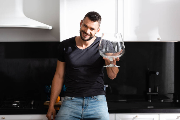 cheerful bearded man holding huge wine glass in kitchen  - Photo, image