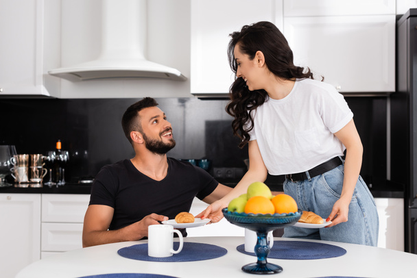 fröhliche Frau hält Teller mit Croissants neben glücklichem Freund in der Küche  - Foto, Bild