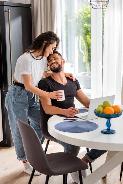 happy girl holding cup near cheerful freelancer boyfriend at home  - Photo, Image