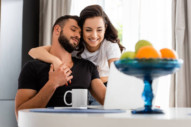selective focus of happy girl hugging handsome boyfriend near laptop, smartphone, cup and fruits  - Photo, Image