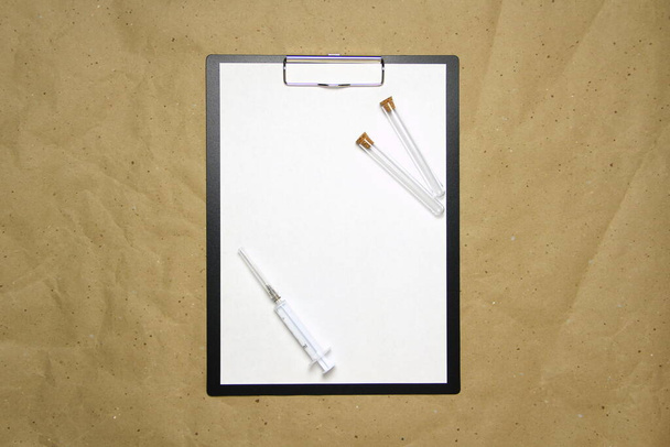 The tablet with a clip for the clip of paper with a white sheet a4 lies against the background of craft brown crumpled paper. On top is a medical syringe and two transparent empty tubes with stoppers. Place for text and layout for design. - Foto, afbeelding