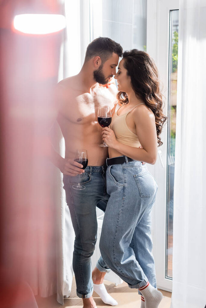 selective focus of muscular man holding glass of red wine near pretty girlfriend in jeans  - Фото, изображение