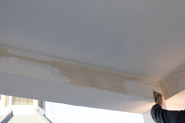 Квалифицированный работник распространяет лепнину и краску на балке террасы во время ремонта квартиры. Белая стена при обработке рабочих, бритье шпаклевкой и покраска
 - Фото, изображение